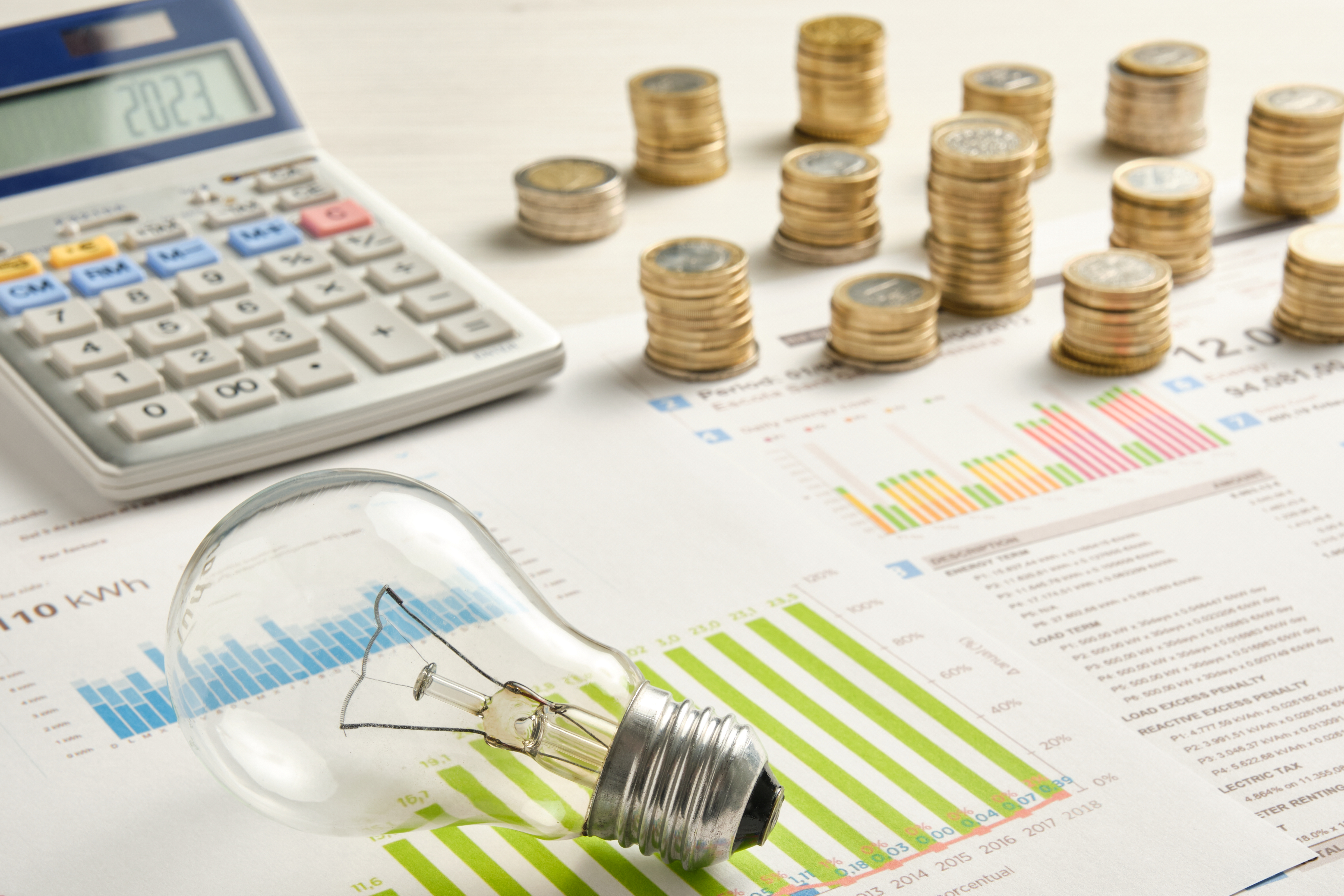 Stacks of coins, a light bulb, calculator & financial charts representing energy costs and savings